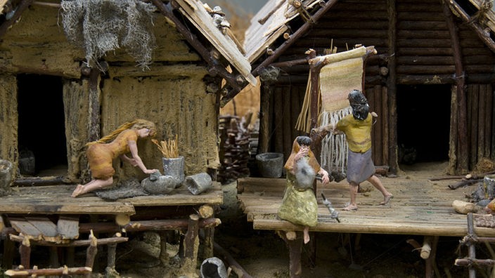 Ein Modell erzählt von der Lebensweise in der Jungsteinzeit. Zwei Holzhütten werden gezeigt, die sogar eine Art terasse - einen Vorbau aus Holz - hatten. Frauen verrichten Hausarbeiten.
