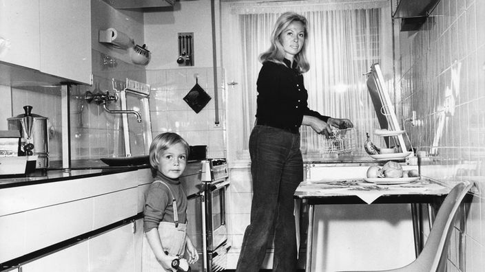 Eine Frau in der Küche, daneben ein kleiner Junge in kurzen Hosen, alles im Stil der 1970er Jahre