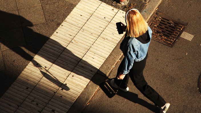 Eine junge Frau trägt Kopfhörer und blickt auf ihr Smartphone, während sie über eine Straße geht.