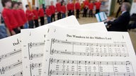Notenblätter zum Lied "Das Wandern ist des Müllers Lust", im Hintergrund steht ein Chor